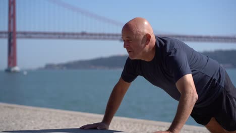 Bald-mature-sportsman-doing-push-ups-at-riverside